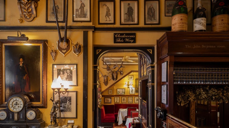 Historic interior featuring framed portraits, decorative antiques, mounted antlers, and a sign reading “Cloakrooms This Way,” with a view through an archway to a red-upholstered dining area.